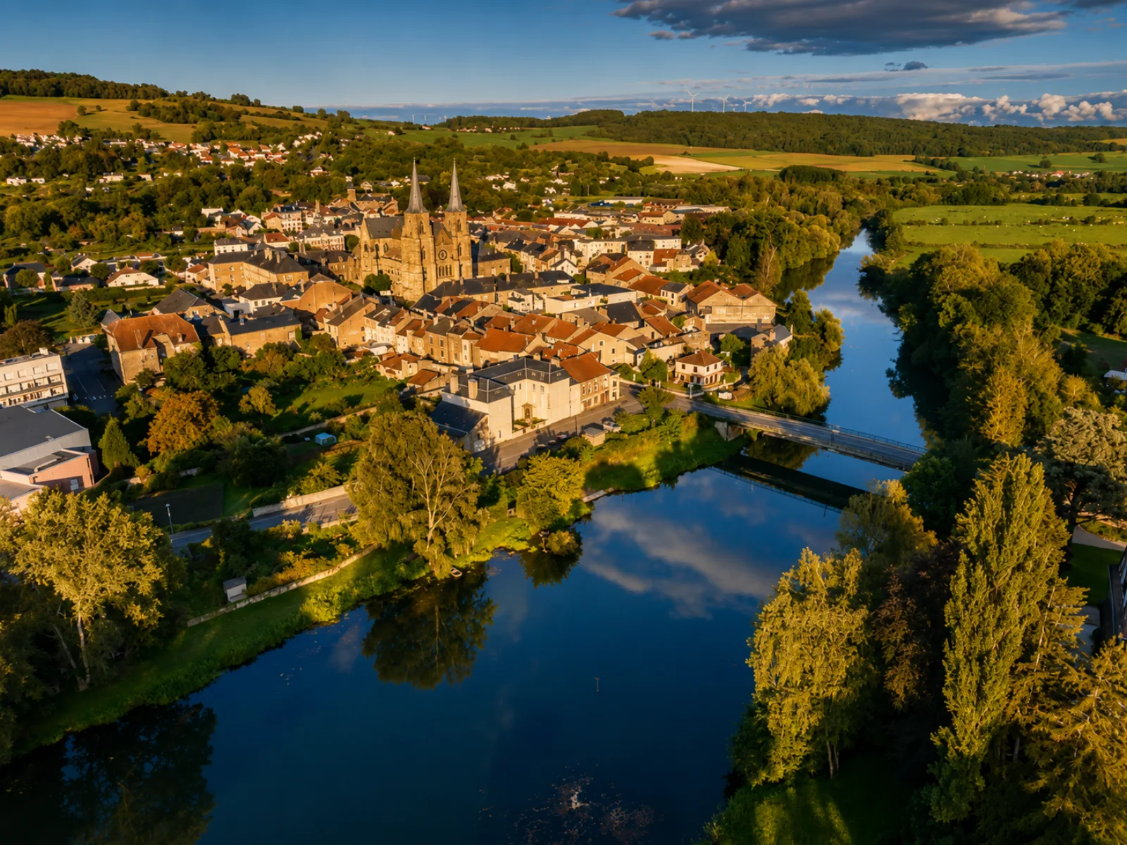 Un bourg ardennais serré autour de son abbatiale à deux flèches, dans un méandre de la Meuse — collines cultivées et forêts du plateau