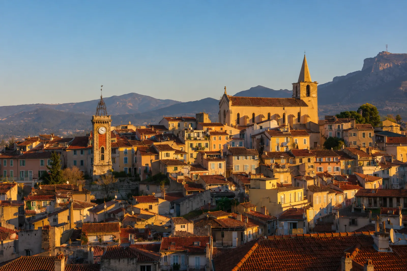Le vieil Aubagne au couchant — toits serrés, campanile de fer et clocher au-dessus de la ville, au pied des reliefs calcaires