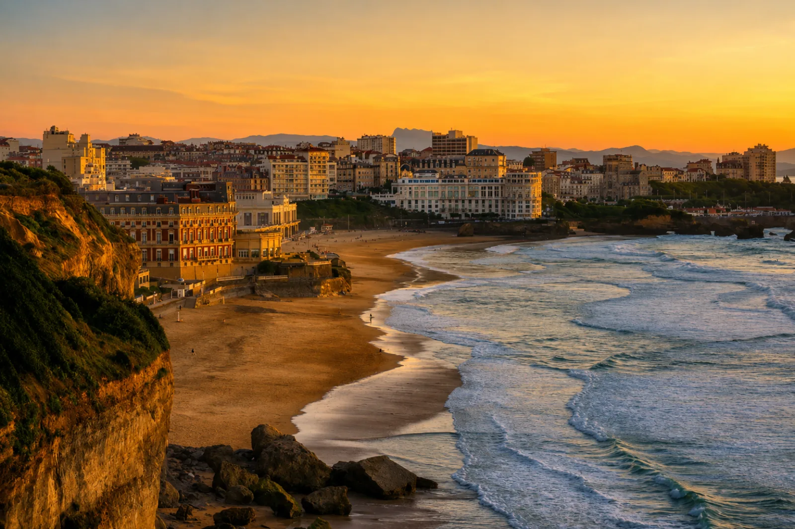 La Grande Plage de Biarritz au couchant, l'Hôtel du Palais sur son rocher et la Rhune à l'horizon — la côte basque entre falaises, houle et ville dense