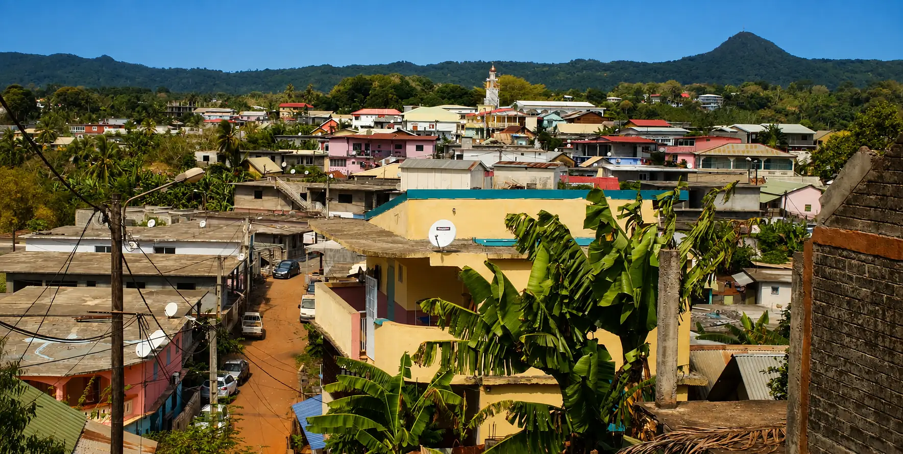 Un village de Mayotte étagé sur ses collines — piste de latérite rouge, toits de tôle et minaret, reliefs boisés en arrière-plan