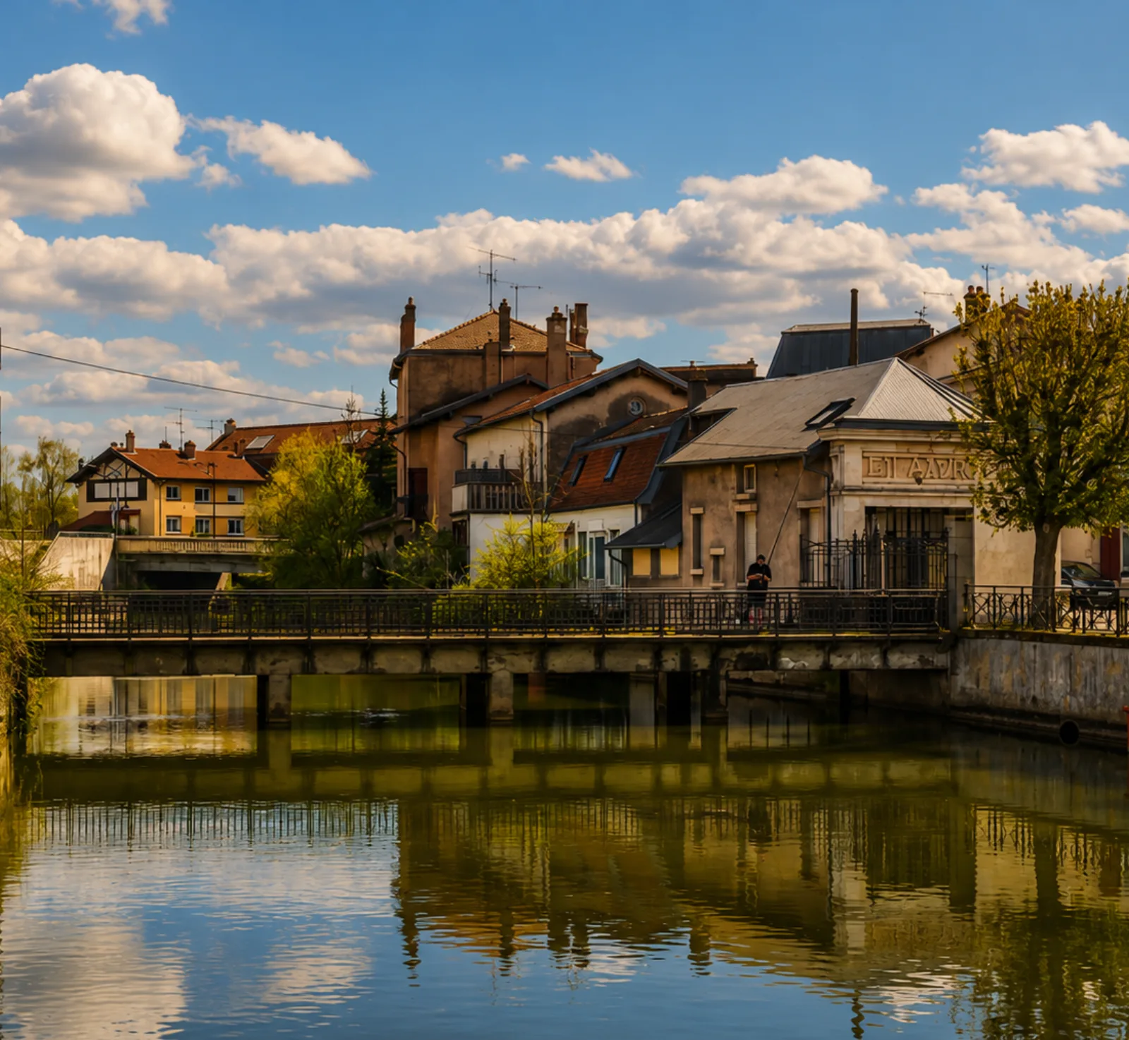 Une passerelle sur le canal, maisons lorraines aux toits pentus serrées sur la berge — en Meurthe-et-Moselle, l'eau borde les cœurs de bourg