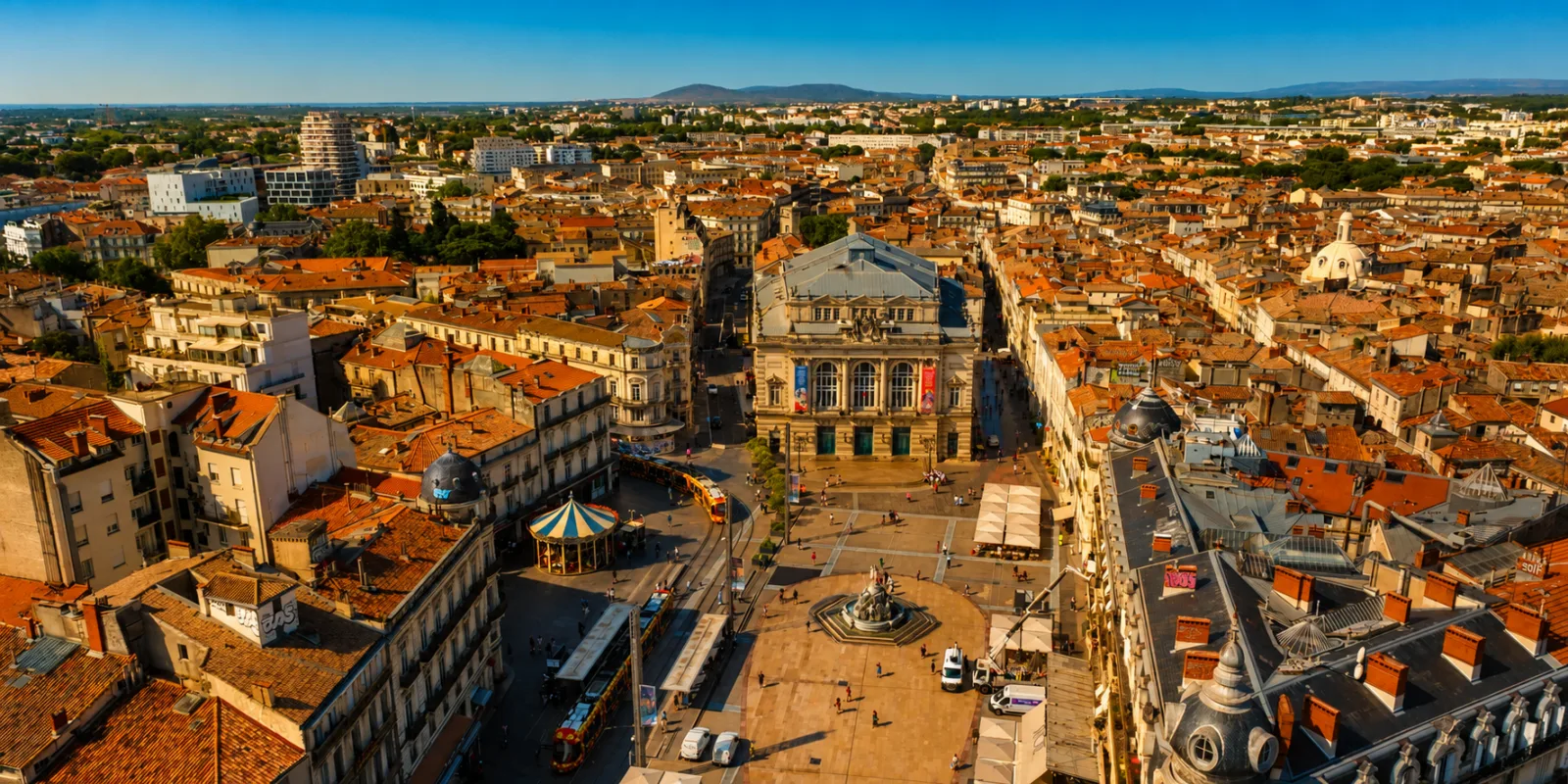 La place de la Comédie et l'Écusson vus d'en haut — les trams de Montpellier dans la ville ancienne, les reliefs de la garrigue à l'horizon