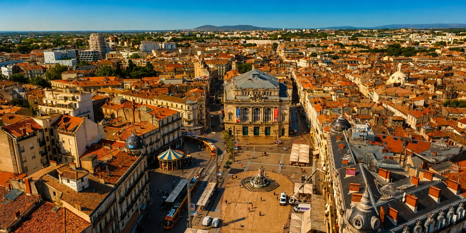 La place de la Comédie et l'Écusson vus d'en haut — les trams de Montpellier dans la ville ancienne, les reliefs de la garrigue à l'horizon