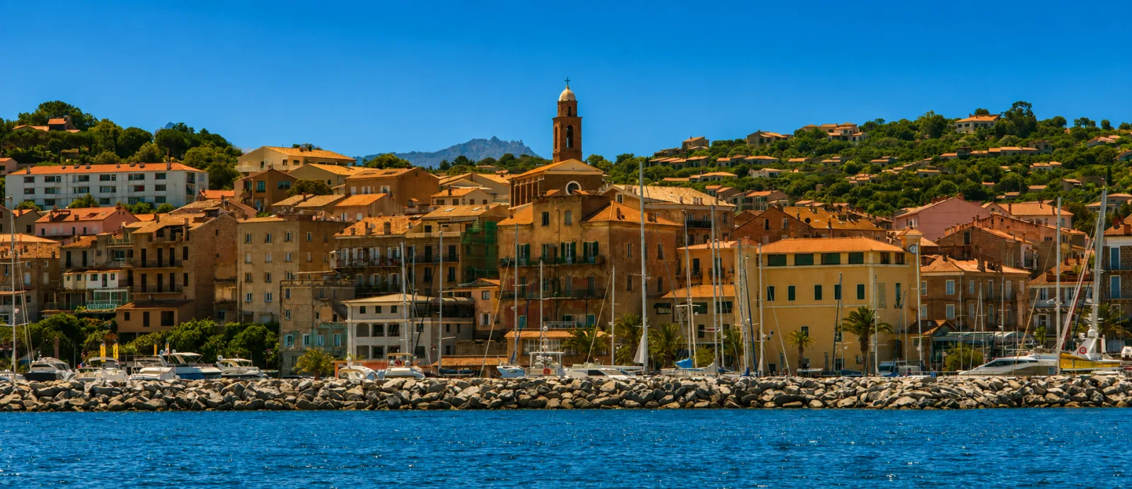 Le front de mer de Propriano vu du large — façades ocre et campanile à coupole au-dessus des quais, derrière la digue en enrochements du port de plaisance