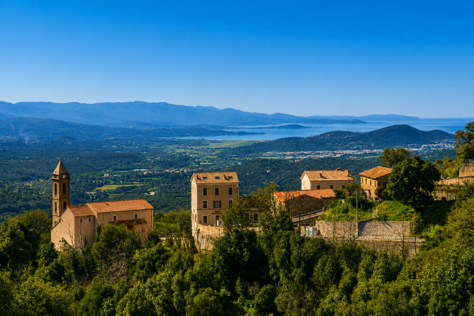 Sarrola-Carcopino et son campanile dominant la basse vallée de la Gravona — la plaine alluviale, l'aéroport et le golfe d'Ajaccio au loin